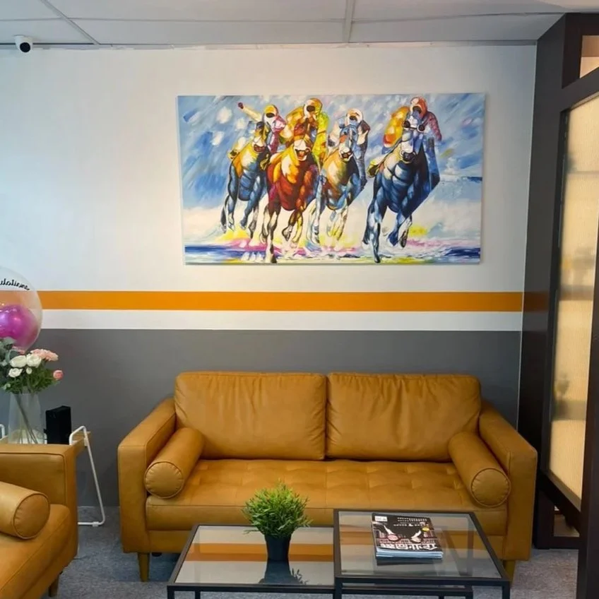 Painting of horse race with five horses and jockeys on a wall above a tan leather sofa in a waiting room or office space, with a glass-topped coffee table, magazine, and potted plant in front.