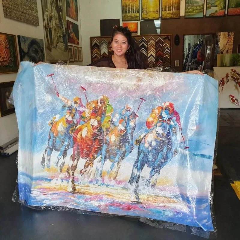 A woman in an art store holding a colorful painting of horse racing, wrapped in plastic.