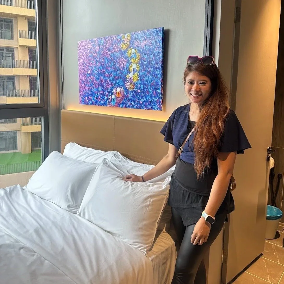 A woman with long, wavy brown hair wearing a black top and black leggings, smiling and standing next to a bed with white pillows and bedding in a modern hotel room. She has sunglasses on her head, a sports watch on her left wrist, and a crossbody bag