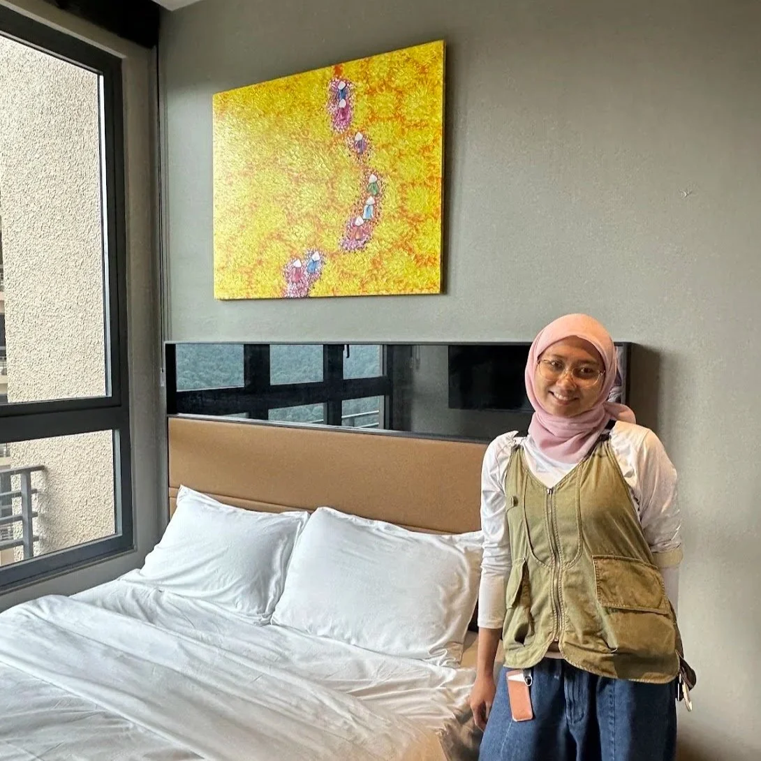 A woman with glasses wearing a pink hijab, white shirt, and tan vest smiling and standing next to a bed in a modern room with gray walls, a colorful abstract painting, a window, and a mirror.