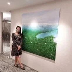 A woman in a dark dress with floral patterns standing next to a large landscape painting of a green field and sky in an indoor setting.