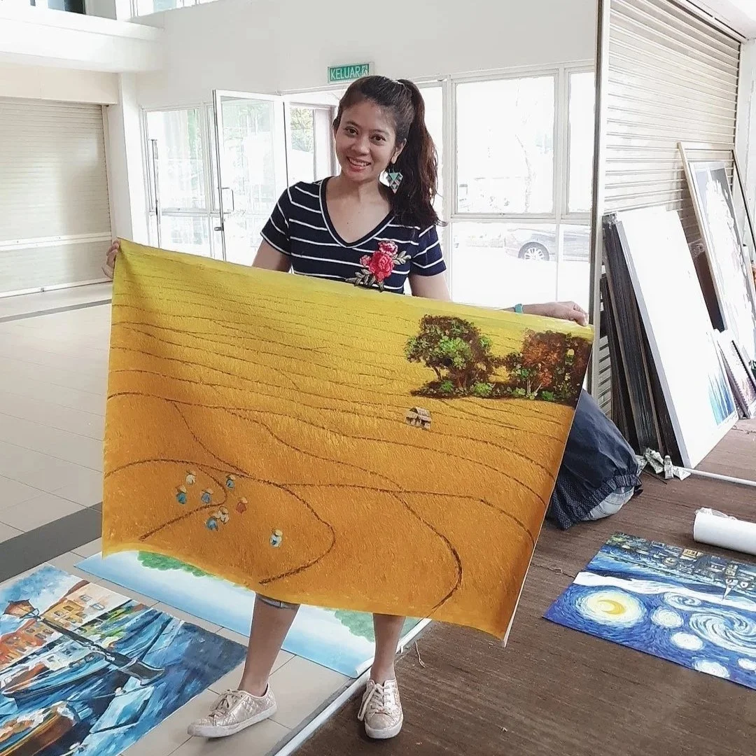 A woman with a ponytail smiling and holding a landscape painting of a yellow field with trees and clouds in the background inside an art gallery.