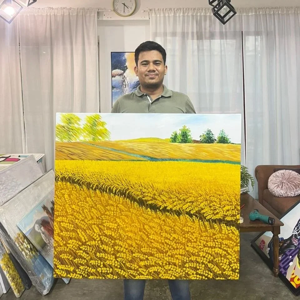 A man holding a large painting of a landscape with yellow fields and green trees in an indoor setting.