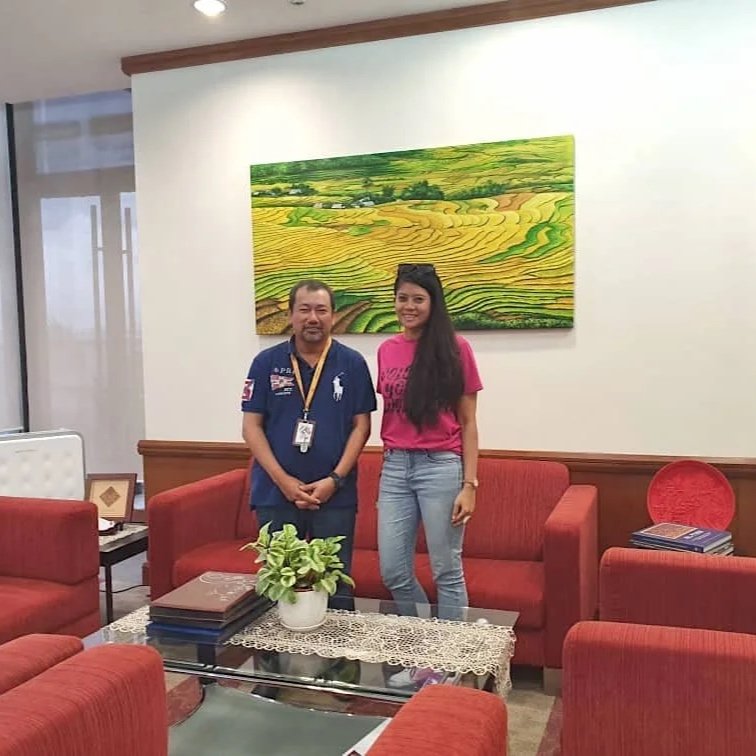 Two people standing in a cozy lounge area with red sofas, a coffee table with a potted plant, and a large landscape painting of green fields hanging on the wall behind them.