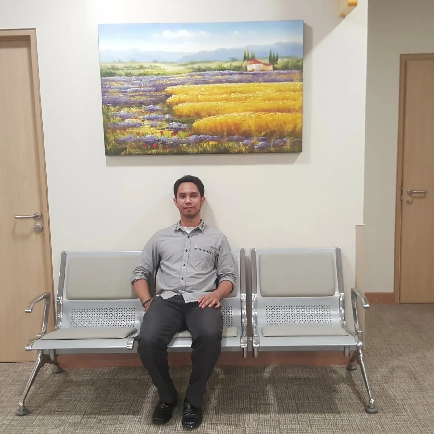 A young man sitting on a bench inside a waiting room with two more empty benches, a large landscape painting on the wall behind him, and closed wooden doors on either side of the painting.