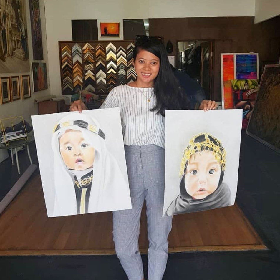 A woman holding two large portrait paintings of children, standing in an art gallery or studio with various artworks and framed pictures displayed on the walls behind her.