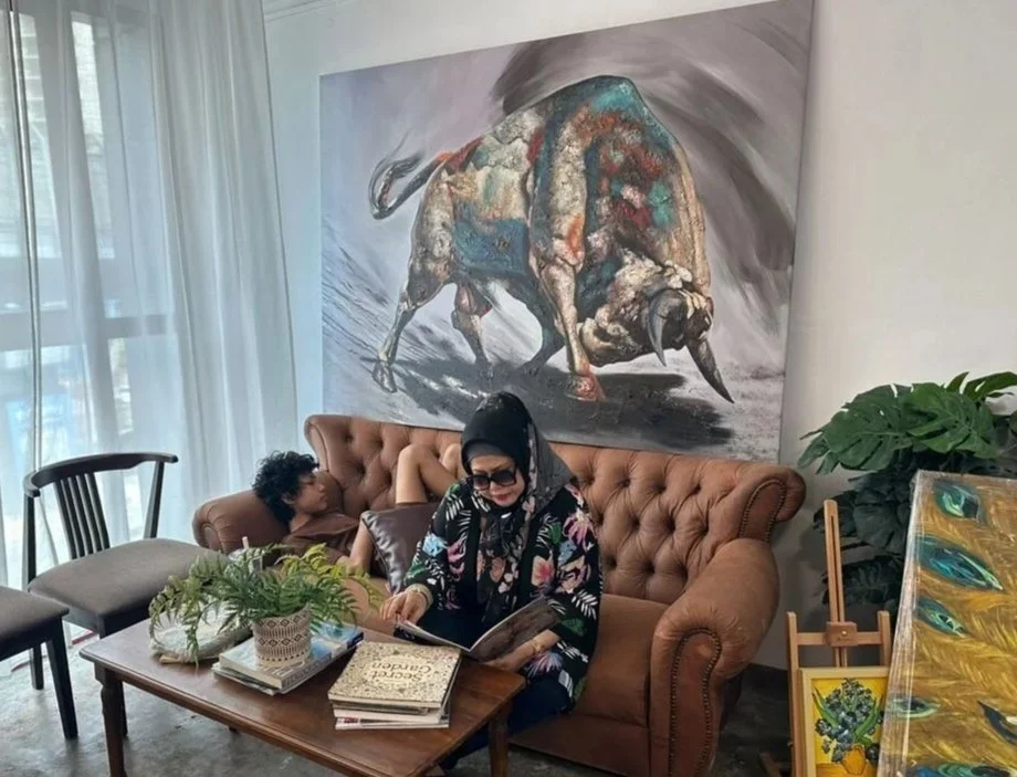A woman in a floral scarf and black headscarf, wearing sunglasses, is sitting on a brown tufted sofa reading a book. A child is lying on the same sofa with their legs up, looking away. There are books and plants on a wooden coffee table in front of t