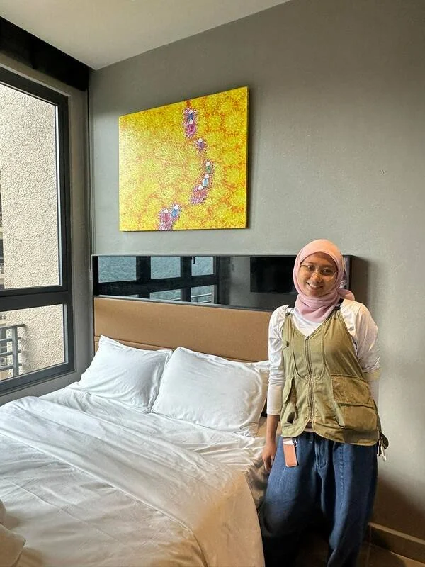 A woman wearing a pink hijab and glasses, smiling, standing next to a bed with white sheets in a modern bedroom. The room has a gray wall, a window, and an abstract yellow artwork hanging above the bed.