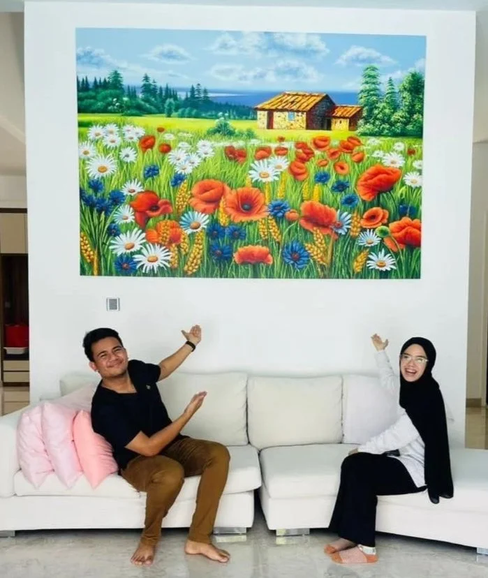 Two people sitting on a white sofa in a living room, smiling and pointing at a colorful landscape painting of a cottage surrounded by a field of flowers, with green trees, blue sky, and distant water in the background.
