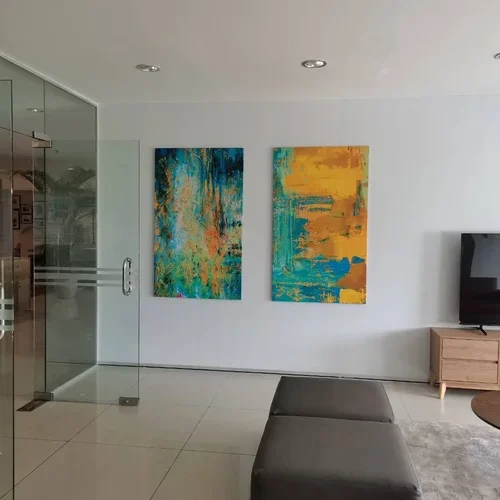 Modern living room with white walls, two colorful abstract paintings hanging side by side, and a television on a wooden stand. There is a black leather ottoman and a glass door on the left.