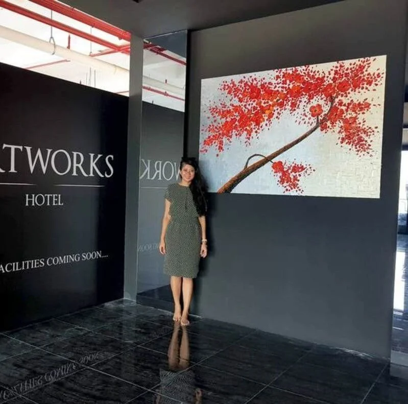 A woman standing inside a hotel lobby near a large wall painting of a tree with red leaves.