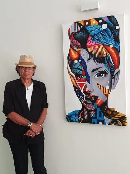 An elderly man wearing a black blazer, white shirt, glasses, and a straw hat standing next to a colorful, abstract portrait of a woman with striking eyes displayed on a white wall.