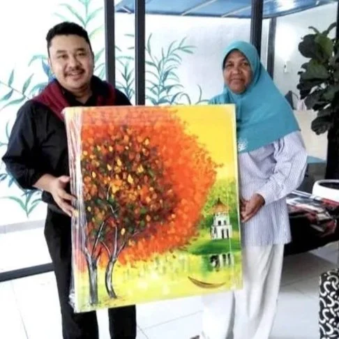 Two people, a man and a woman, standing indoors holding a large painting of a colorful tree with an abstract background and a small house near a pond.
