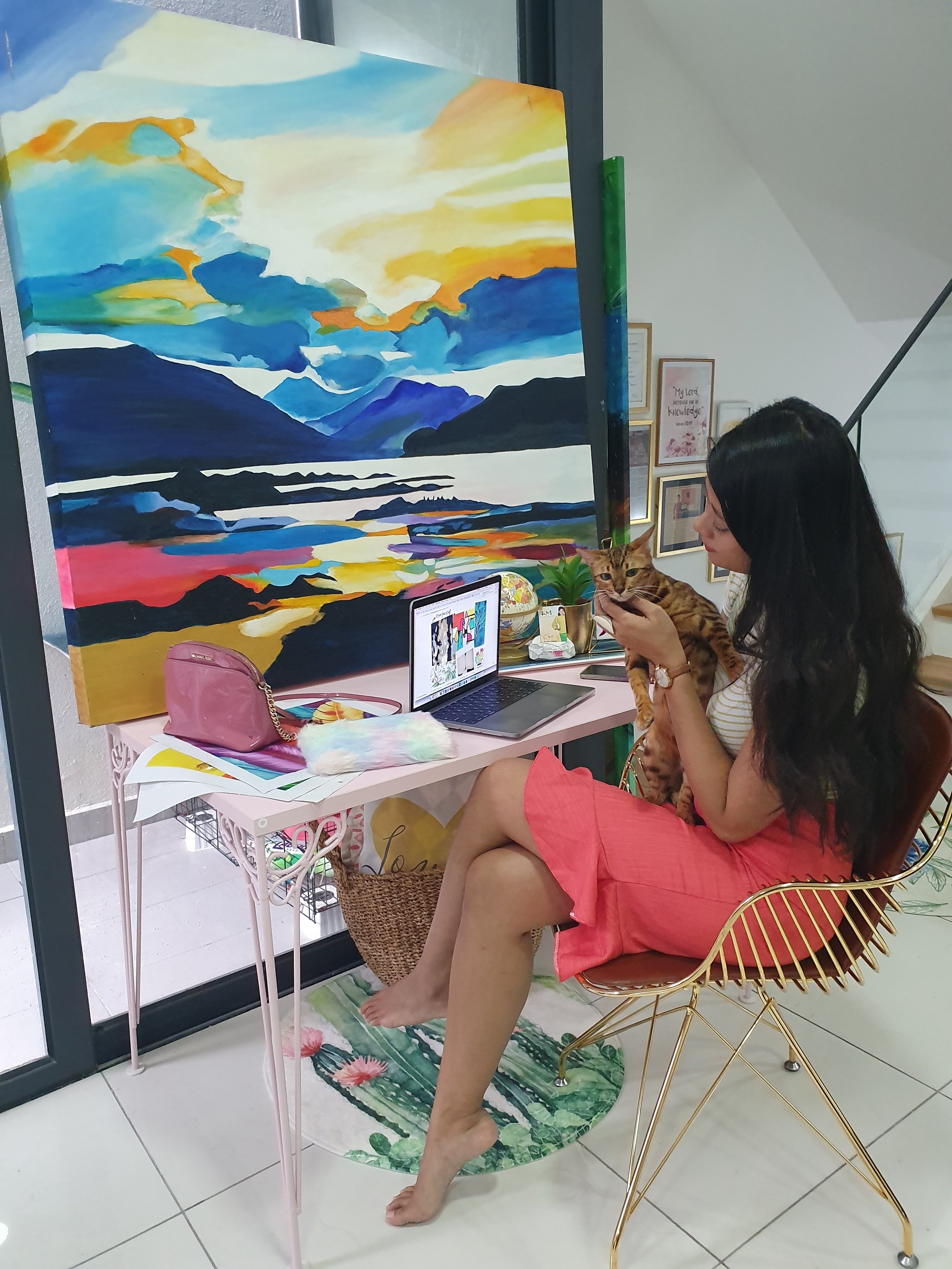 A woman sitting on a gold wire chair, holding a cat, next to a pink table with a laptop, a bag, and colorful items, in front of a large, abstract landscape painting with mountains and a sunset, in a well-lit room.