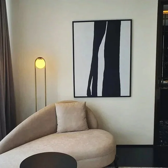 A beige velvet curved sofa with a single beige pillow, a black round side table, a tall floor lamp with a yellow glow, and an abstract black and white painting on a light-colored wall.