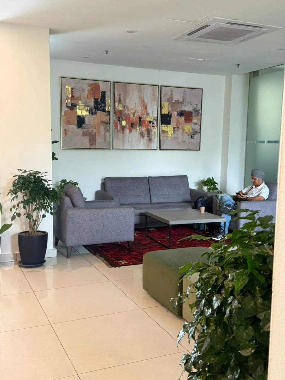 Modern hotel lobby or lounge area with gray sofas, a small mirror table, artwork on the wall, potted plants, and a man sitting on a sofa using his phone.