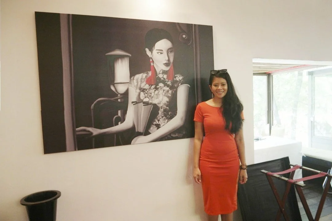 Woman in a red dress standing next to a large black-and-white portrait of an Asian woman with earrings and a floral dress, in an indoor space with windows and furniture.