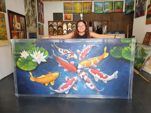 A woman with glasses and long dark hair smiling and extending her arms in front of a colorful koi fish painting at an art gallery, surrounded by various framed artworks and landscape paintings.