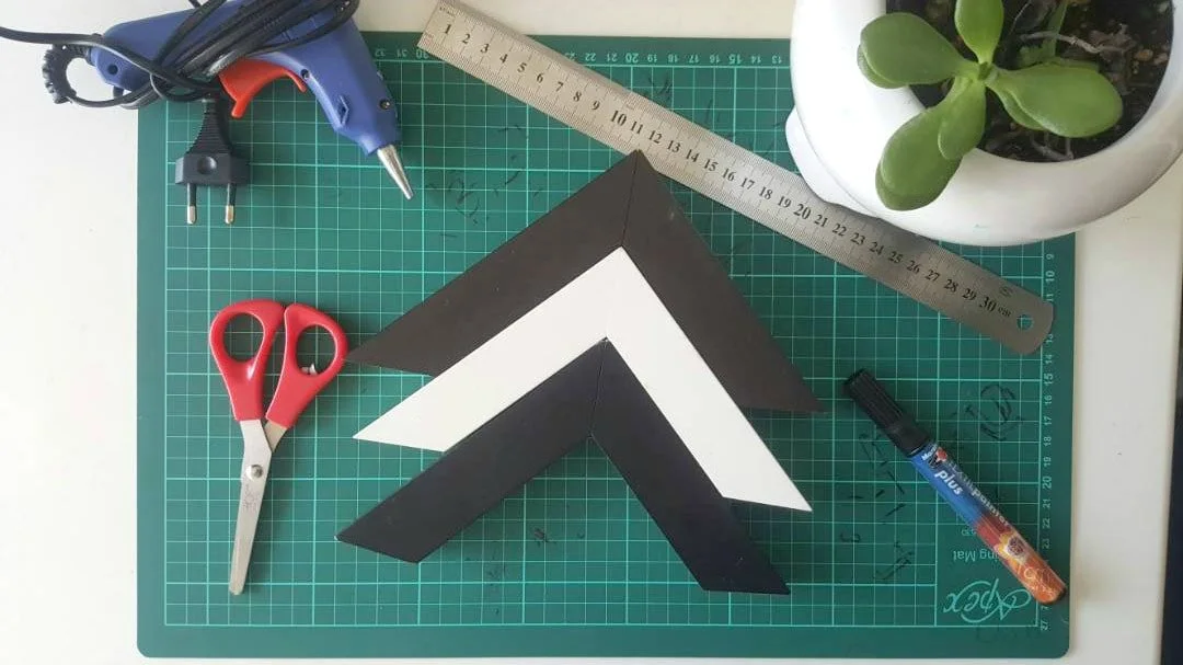 Top view of a workspace with a green cutting mat, black and white paper chevron shapes, a blue hot glue gun, red scissors, a metal ruler, a black marker, and a potted plant with green succulent leaves.