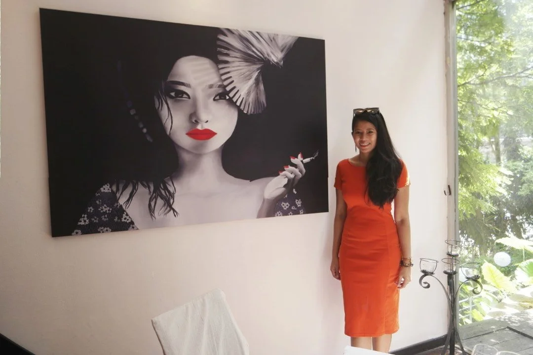 A woman in an orange dress standing beside a large black and white portrait of a woman with lips painted red, holding a cigarette, with a decorative fan in her hair.