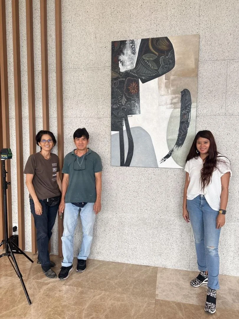 Three women standing beside a large abstract painting on a wall, with a wooden slat decor on the left.