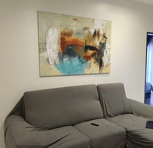 A gray fabric sofa with a remote control resting on it in a living room. Above the sofa is a large abstract painting with various colors and shapes. There's a doorway on the right side leading to a room with a bed.