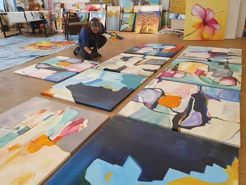 Artist arranging large abstract and floral paintings on the floor in an art studio.