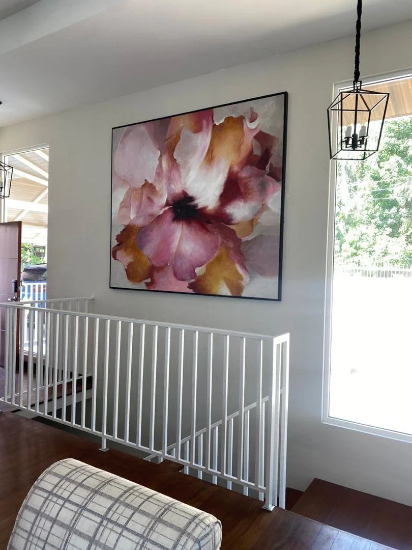 Interior of a home with a large abstract floral painting on the wall, white railing by the staircase, a window letting in natural light, and a plaid-patterned cushion in the foreground.