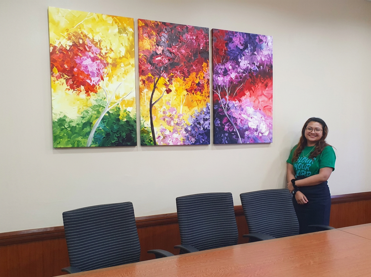 A woman in a green shirt and glasses standing in a conference room next to a large colorful painting of trees with red, purple, yellow, and green leaves.