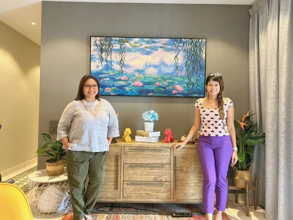 Two women standing in a living room in front of a wooden sideboard with decorative books and flowers, a framed impressionist-style painting on the wall behind them, and potted plants on either side.
