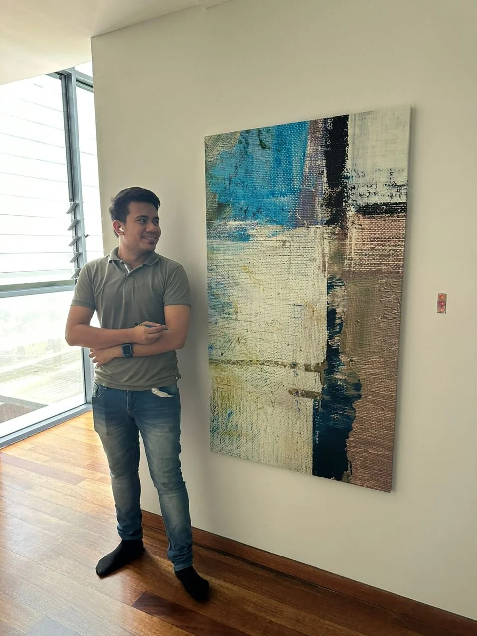 Young man in gray T-shirt and jeans standing indoors next to an abstract painting on a white wall, with large windows in the background.