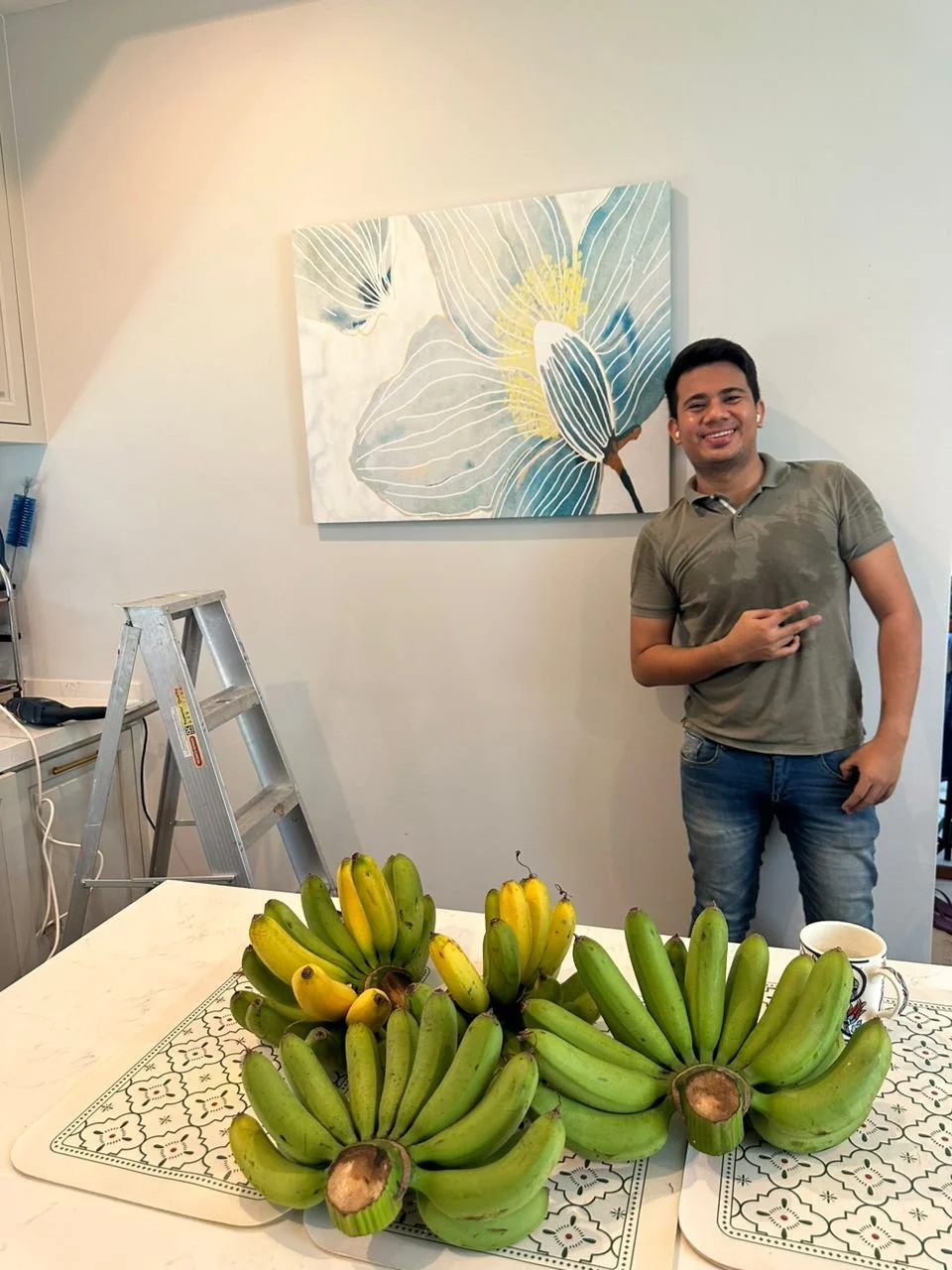 A man smiling and making a peace sign stands next to a table with bunches of bananas. Behind him, a floral painting decorates the white wall, with a ladder and some tools nearby.