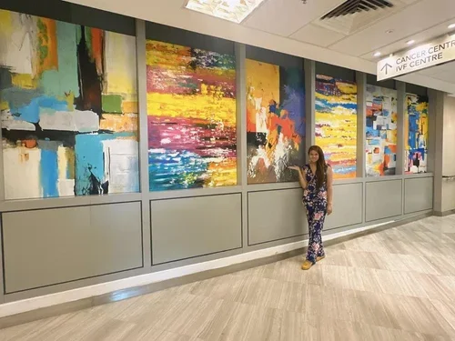 A woman standing beside colorful abstract paintings in a hallway with a sign pointing to a cancer treatment center.