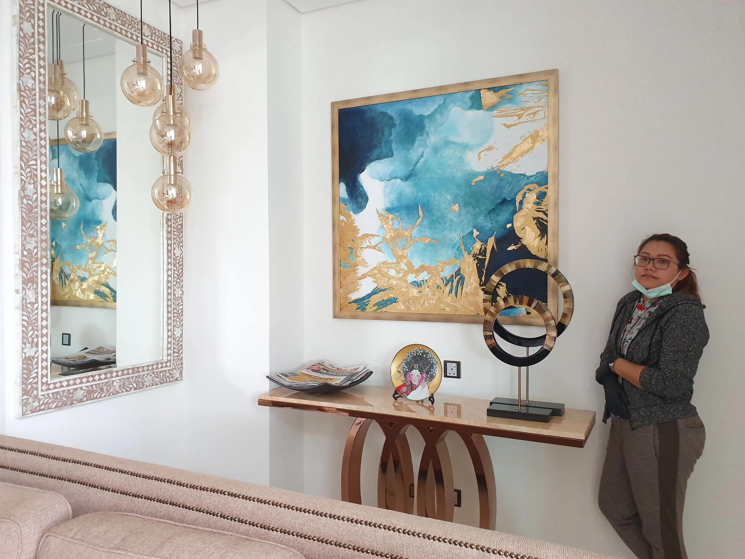 A woman wearing glasses and a face mask stands next to a wooden table displaying decorative items including a circular mirror with a gold and black frame, a small decorative plate with an illustration of a woman in traditional dress, and two modern a