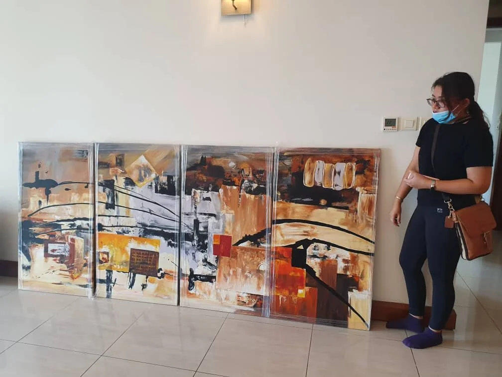 A woman wearing a face mask, glasses, black t-shirt, dark pants, and purple slippers, standing next to abstract paintings leaning against a wall in an indoor setting.