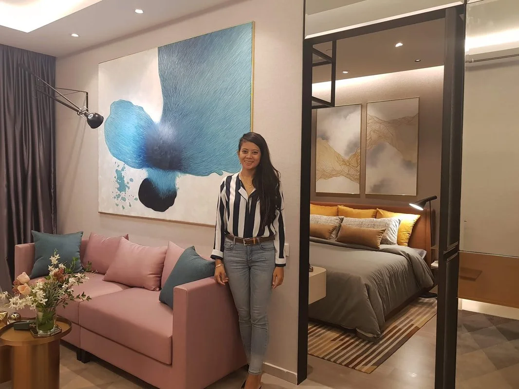 A woman with long dark hair standing in a modern living room, smiling, wearing a black and white striped shirt and jeans. Behind her is a pink sofa with teal and pink pillows, a large abstract blue and black painting on the wall, and a bedroom visible through a black metal frame divider with a bed, decorative pillows, and gold artwork.