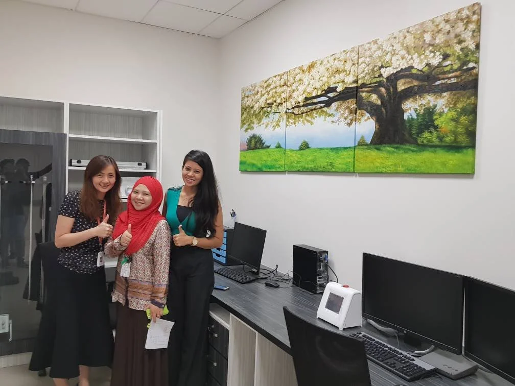 Three women standing in an office next to computer desks; one in a polka dot shirt, another in a red hijab and patterned blouse, and the third in a black and teal dress. All are smiling and giving thumbs up, with a large framed painting of a flowerin