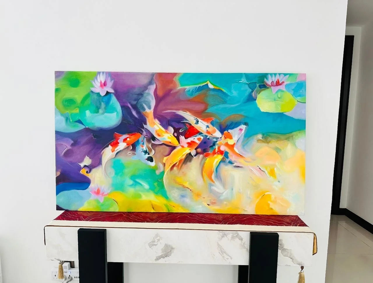 Colorful abstract painting of koi fish swimming among water lilies, displayed on a white wall with a white marble stand.