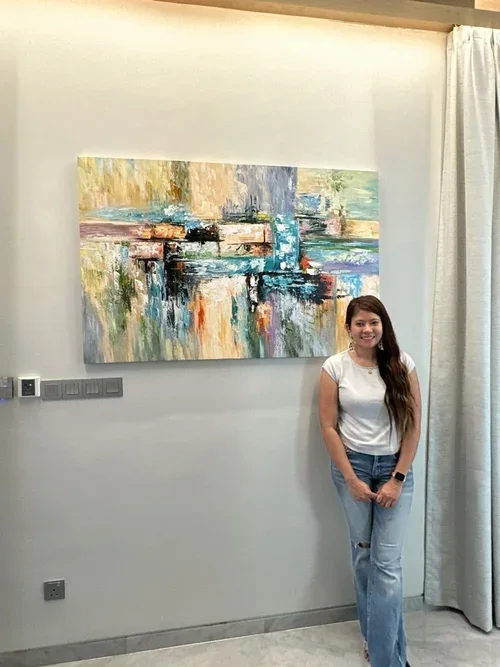 A young woman with long dark hair, wearing a white sleeveless shirt and ripped jeans, standing next to an abstract painting on a white wall, with a curtain on the right side.