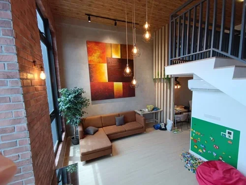 Living room with brick wall, large window, modern artwork, hanging lights, brown sofa, plants, and a play area with a small green chalkboard and colorful toys.