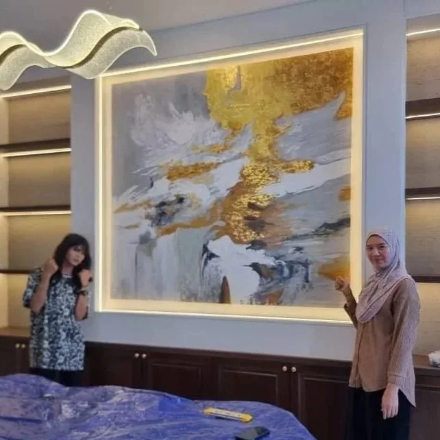 Two women standing on either side of a large abstract painting with gold, white, and black accents in a modern interior space with wooden shelves and a blue table in the foreground.
