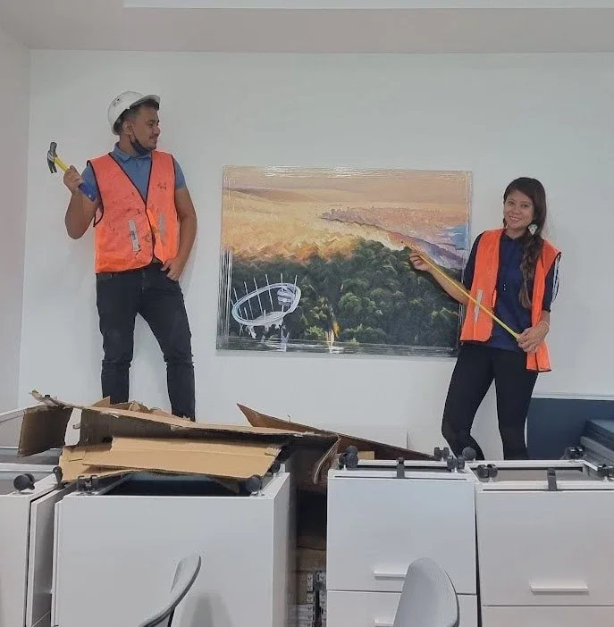 Two people wearing orange safety vests, with one holding a hammer and the other a measuring tape, standing in front of a painting of a landscape with trees and a body of water. There is a partially opened cardboard box on a desk in the foreground.
