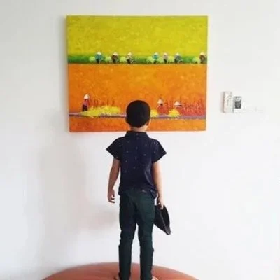 A boy with dark hair, wearing a navy shirt and jeans, stands in front of a colorful abstract painting on a white wall. The painting features three horizontal sections in green, orange, and yellow with various small shapes and figures.