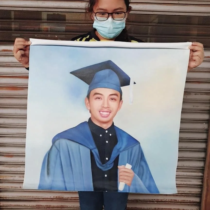 A person wearing glasses and a face mask holding a large portrait of a graduate in a cap and gown.