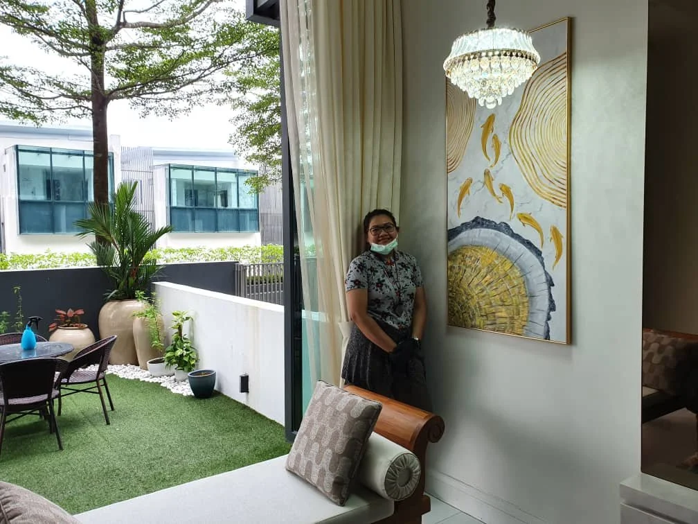 A woman standing inside near a large wall art piece, with a balcony area outside featuring plants and outdoor furniture.