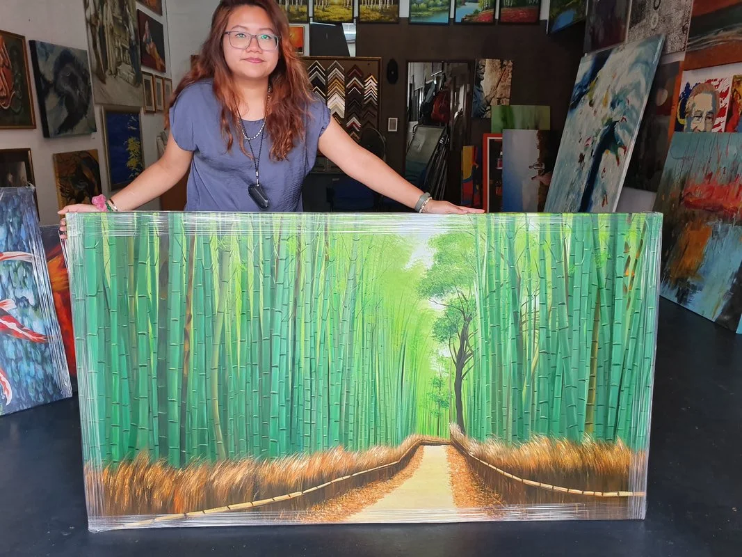 A woman with long reddish-brown hair and glasses stands behind a large painting of a bamboo forest with a pathway, inside an art gallery.