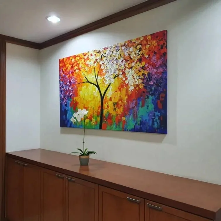 Colorful abstract painting of a tree with orange, yellow, purple, and blue foliage hanging above a brown cabinet, with a potted orchid on top.