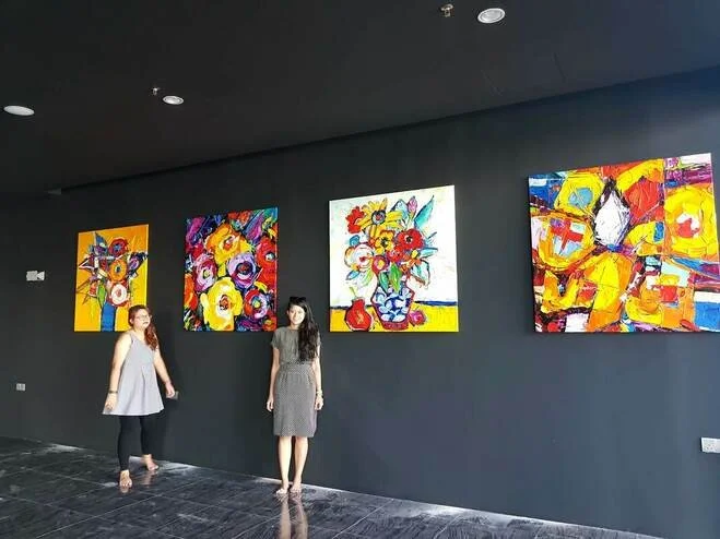 Two women standing in an art gallery with four colorful abstract artwork paintings on a dark gray wall.