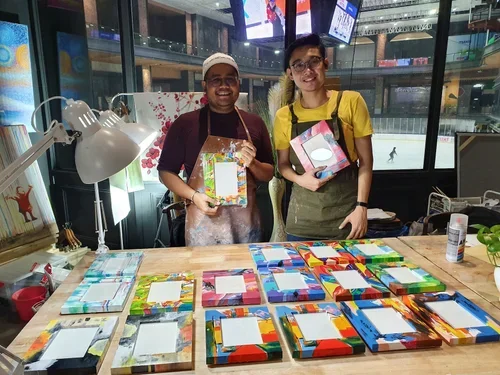 Two people standing behind a table displaying colorful handmade picture frames, with one person holding a frame and the other holding a gift box, inside a crafts workshop.