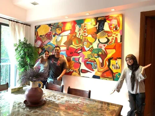 Three people standing in a dining room, smiling, next to a colorful abstract painting on the wall. The room has a large window with off-white curtains, a wooden dining table with a vase of lavender, and a potted plant in the corner.